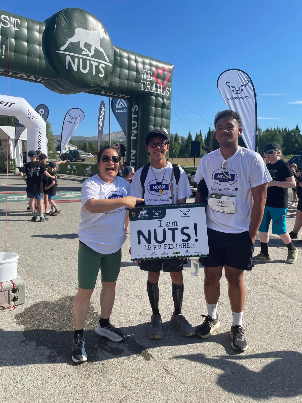 Kolme ihmistä hymyilee ja seisoo tapahtuma-alueella, keskimmäinen pitää kylttiä, jossa lukee 'I am NUTS! 15 KM FINISHER'.
