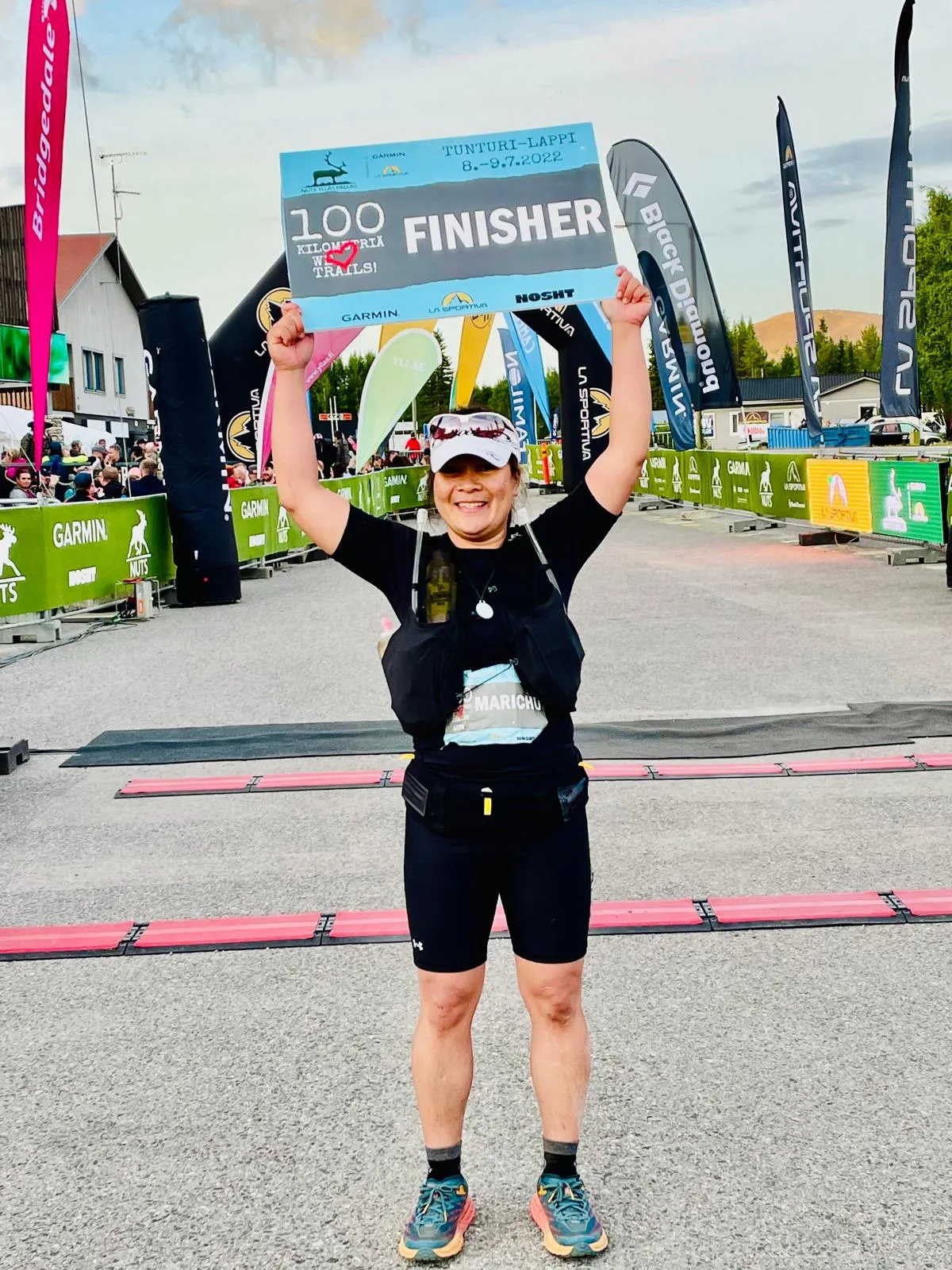 Ilmoitusmerkkejä pitelevä hymyilevä juoksija maaliviivalla Tunturi-Lappi 100 kilometrin polkujuoksukilpailussa.