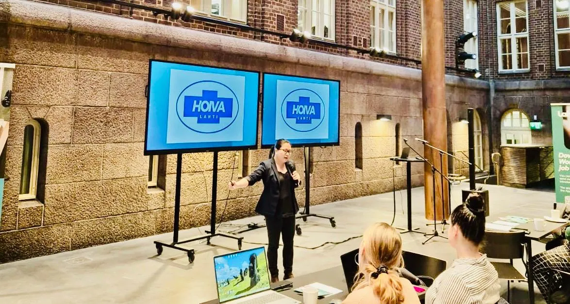 Nainen puhumassa mikrofonin kanssa yleisölle, taustalla kaksi näyttöä, joissa näkyy HOVA Lahti -logo, sisätilassa teollisessa luentosalissa.