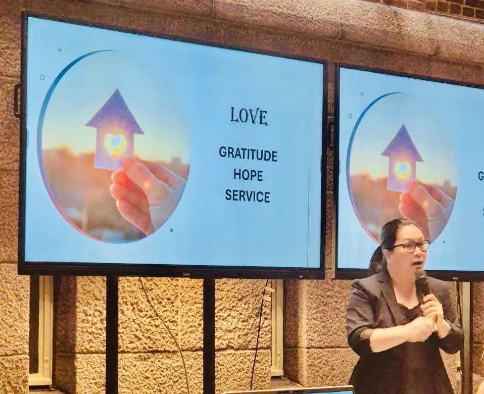 Nainen puhuu mikrofonin kanssa taustalla kaksi näyttöä, joissa kuva kädestä pitämässä talon muotoista esinettä sydänleikkauksella, teksti: RAKKAUS, KIITOLLISUUS, TOIVO, PALVELU.